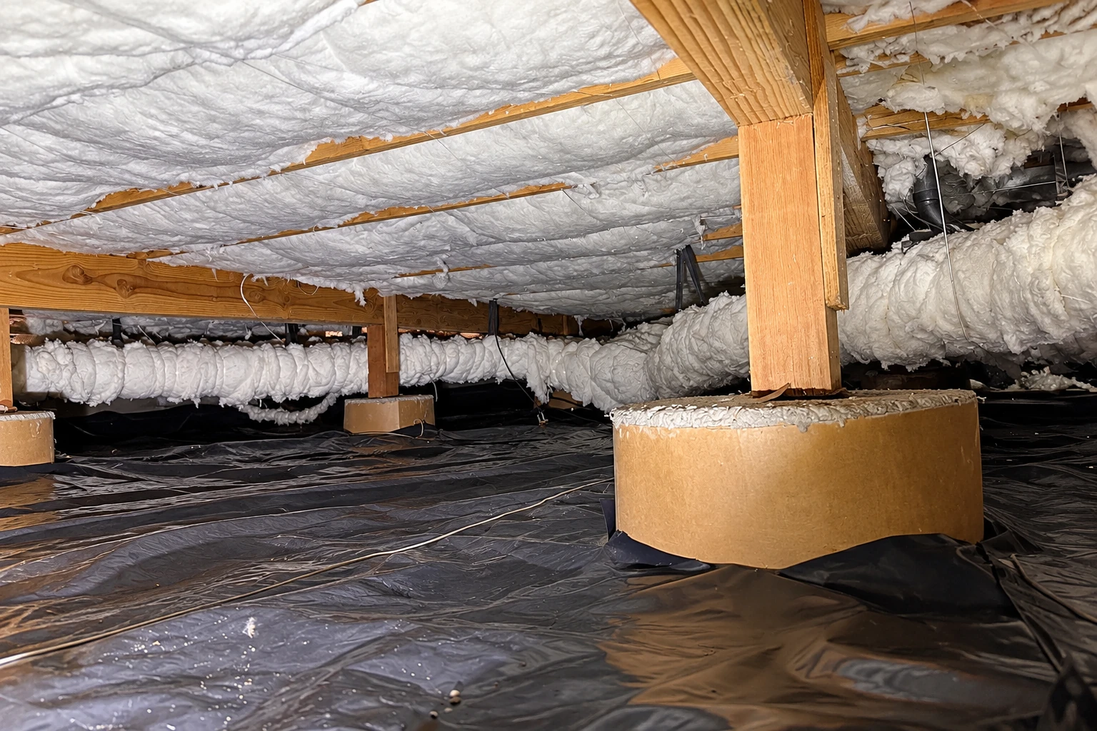 A crawl space has white insulation beneath the joists and a black vapor barrier across the ground.