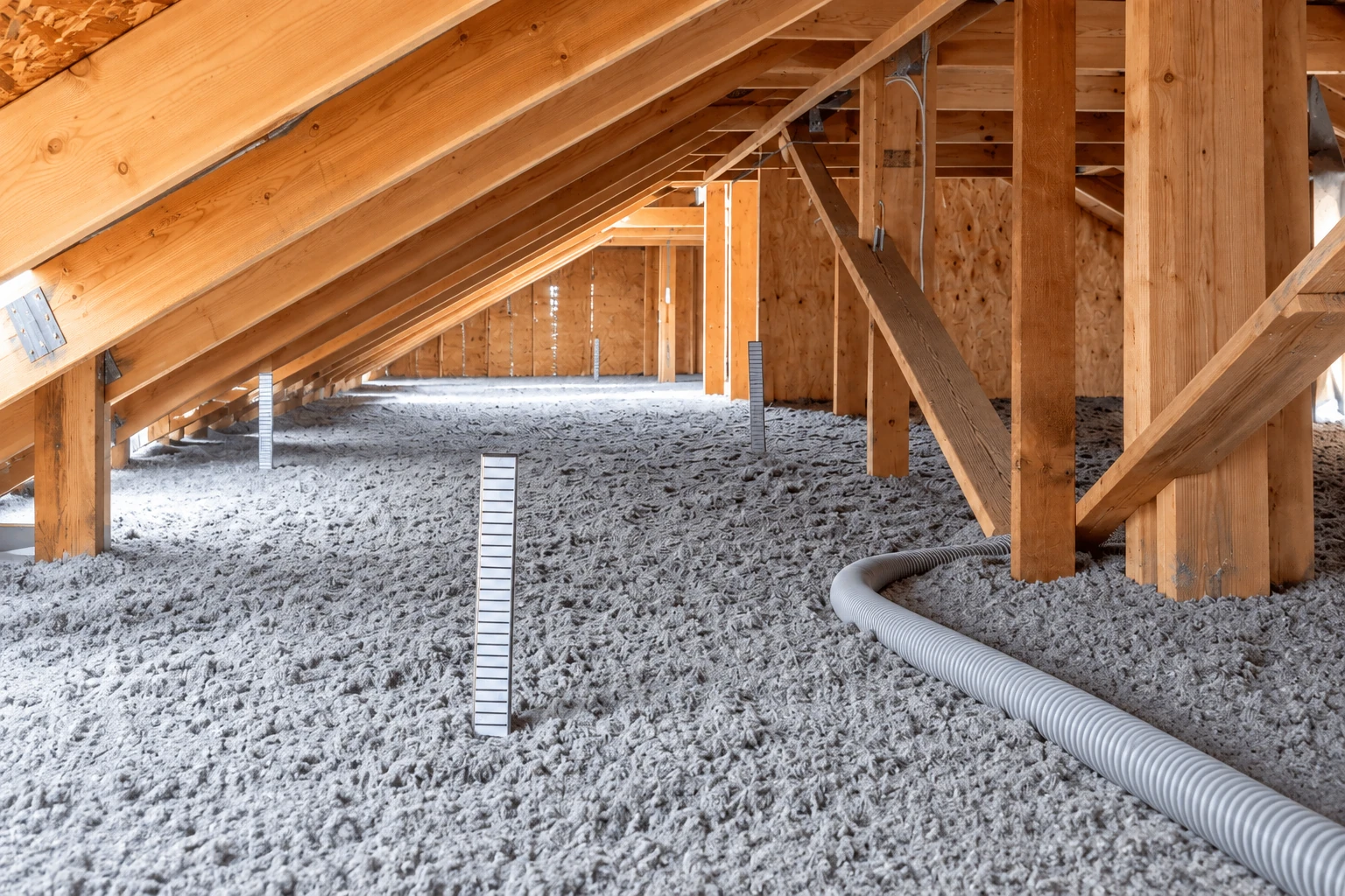 Loose-fill insulation covers an attic floor beneath exposed wooden rafters, with a flexible hose resting across it.