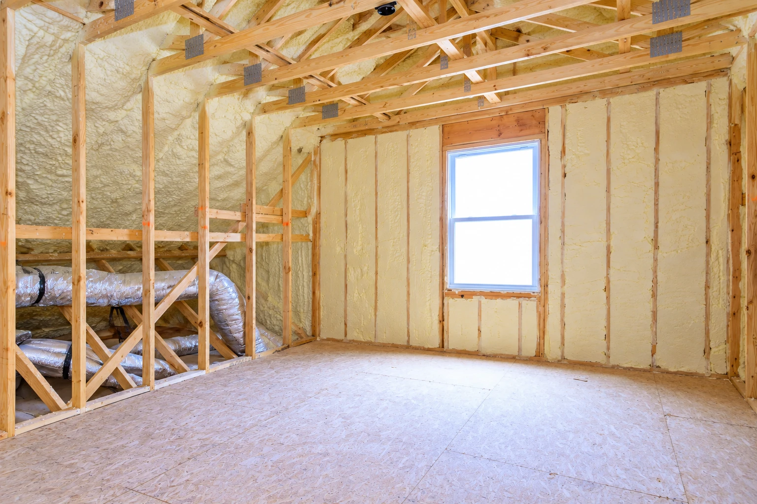 Attic with exposed wooden framing, spray foam insulation on the walls and roof, a window, and unfinished plywood flooring.