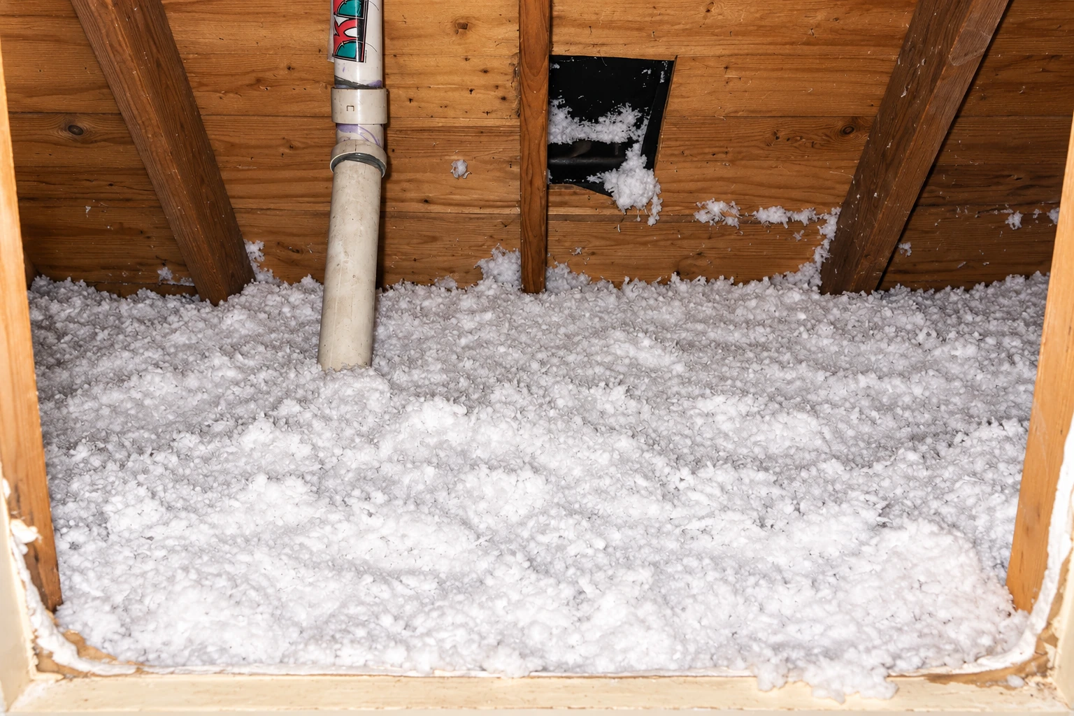Loose white blown-in insulation blankets an attic floor beneath exposed wooden roof boards and a plumbing vent.