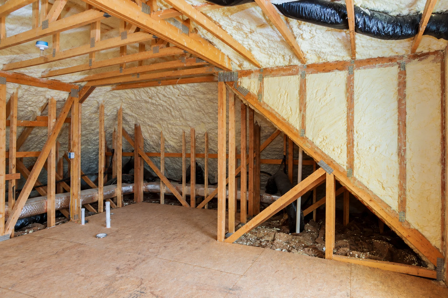 Attic framing with uneven spray foam covering the roof and walls, black ducts overhead, and exposed floor framing.